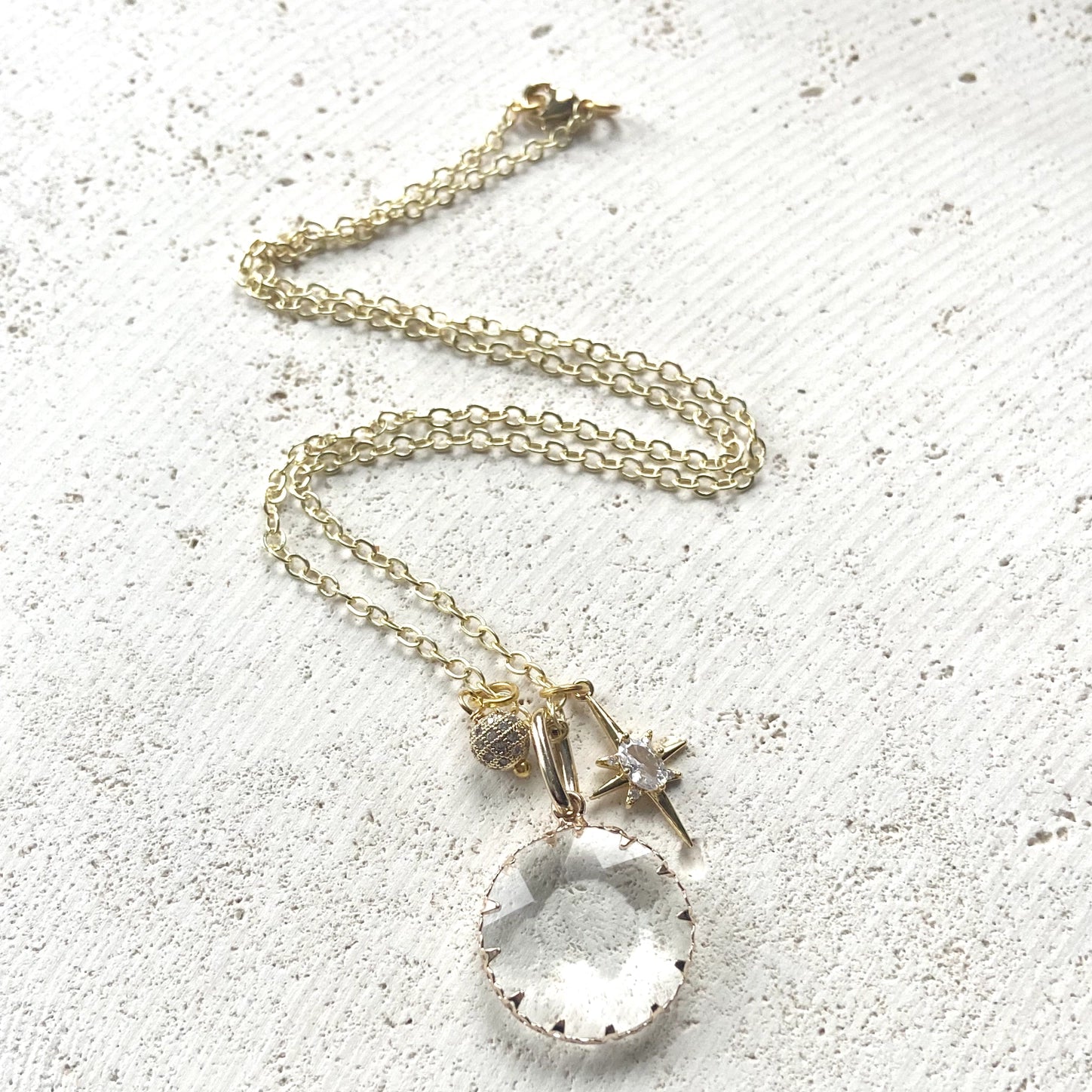 Gold necklace layering a faceted clear crystal pendant, a starburst charm, and a small pavé bead.